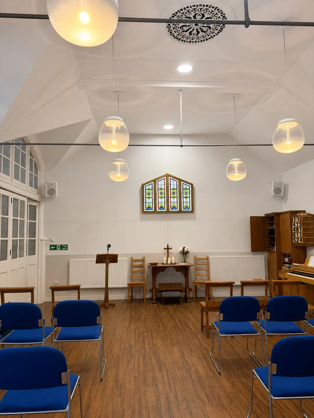The church room facing the altar table, with rows of blue chairs and pendant lighting
