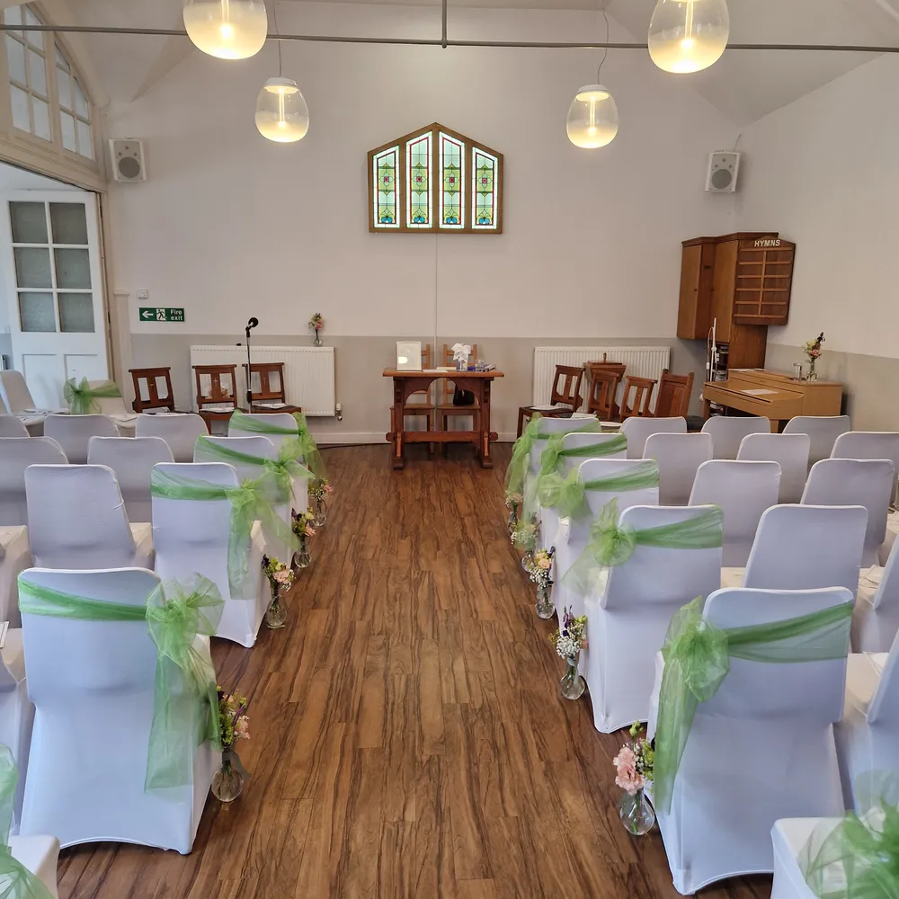 The main hall dressed for a wedding, with white chair covers, green sashes and a centre aisle leading to the altar